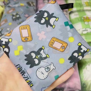 May include: A grey reusable shopping bag with a black and white penguin design. The bag features a repeating pattern of penguins, game consoles, and plus signs. The text "Badtz-Maru" is printed on the bag.