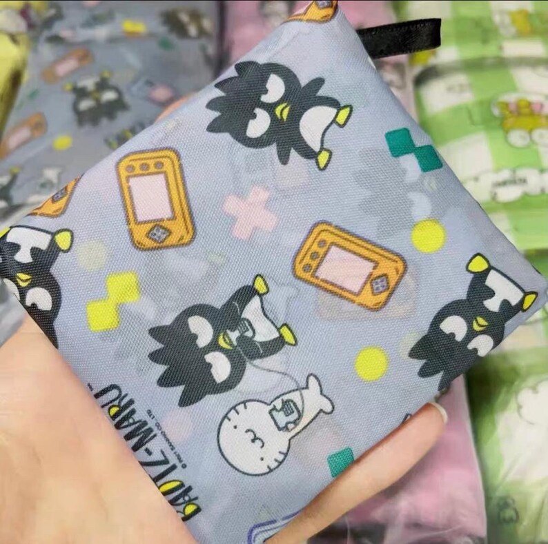 May include: A grey reusable shopping bag with a black and white penguin design. The bag features a repeating pattern of penguins, game consoles, and plus signs. The text "Badtz-Maru" is printed on the bag.