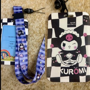 Op de afbeelding: Een zwart-wit geblokte ID-kaart met een cartoonfiguur dat een dienstmeidenkostuum en een schedelband draagt. Het personage heet Kuromi en houdt een roze kers in haar hand. De tekst "Kuromi cut cherry" staat op de kaart gedrukt. Een blauw-witte lanyard met een geruit patroon en een cartoonfiguurontwerp is ook inbegrepen.