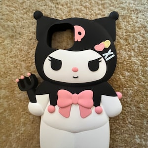 Op de afbeelding: Zwarte en witte telefoonhoes met een roze strik en een cartoonfiguur met een schedel en gekruiste beenderen. Het figuur heeft roze wangen en gele hartjes op zijn hoofd. De hoes is gemaakt van siliconen.