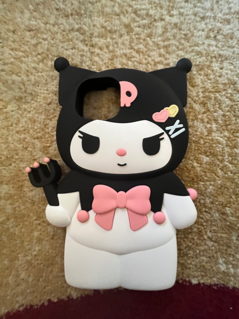 Op de afbeelding: Zwarte en witte telefoonhoes met een roze strik en een cartoonfiguur met een schedel en gekruiste beenderen. Het figuur heeft roze wangen en gele hartjes op zijn hoofd. De hoes is gemaakt van siliconen.