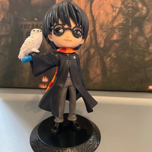 May include: A black and grey Harry Potter figurine with a white owl perched on his arm. The figurine is standing on a black base with a decorative design.