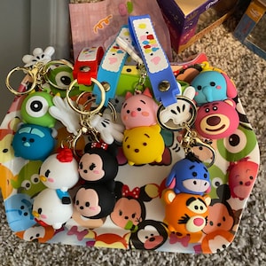 Puede incluir: Una colección de llaveros de personajes coloridos con personajes populares de Disney. Cada llavero tiene un anillo dorado y una correa en varios colores. Los llaveros se exhiben en una bandeja con un diseño de dibujos animados.