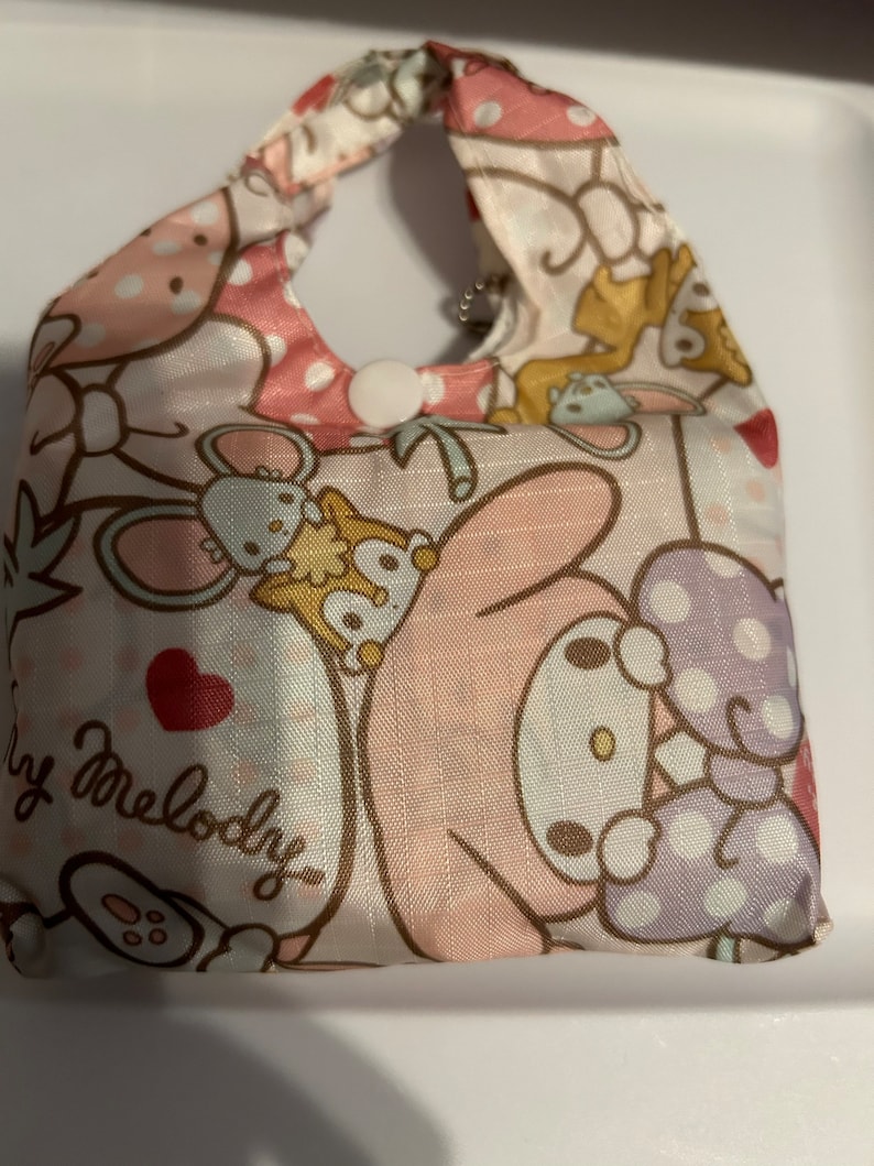May include: A pink and white reusable shopping bag featuring a My Melody character design. The bag has a pink and white polka dot pattern and a white button closure.