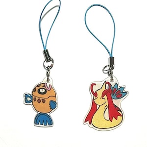 Puede incluir: Dos amuletos de teléfono con personajes de dibujos animados coloridos. El primer amuleto es un pez marrón y azul con un ojo rosa y blanco. El segundo amuleto es una criatura amarilla y roja con un ojo rosa y alas azules.