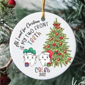 May include: White round Christmas ornament with a cartoon image of two teeth wearing Santa hats and elf hats. The text on the ornament reads "All I want for Christmas is my two front teeth" and "Caleb 2022".