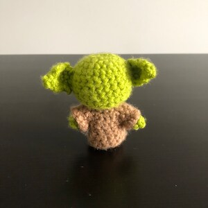 May include: A green and brown crocheted plush toy resembling a character from the Star Wars franchise. The toy is standing on a black surface.