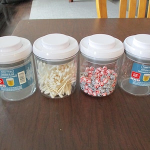 May include: Four clear plastic containers with white lids are arranged on a brown table. Two containers are empty, one is filled with cotton swabs, and another with red and silver beads. The containers have blue and red labels.