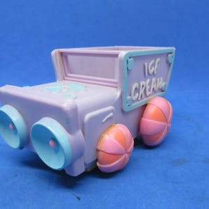 May include: A pastel purple toy ice cream truck with light blue accents. The truck has round blue headlights and pink and orange striped wheels. The sign on the side reads "ICE CREAM".
