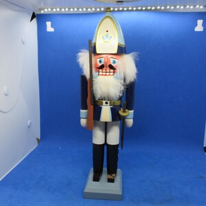 May include: A wooden nutcracker soldier figurine, standing on a gray base. The figurine is dressed in a blue uniform with white accents, a tall hat, and a white beard. It holds a rifle and a sword.