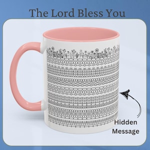 May include: White ceramic mug with a pink handle and interior. The mug features a black and white patterned design with the text "The Lord Bless You" at the top. A hidden message is indicated with an arrow.