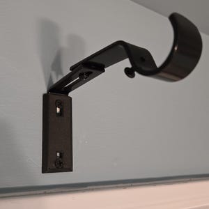 May include: Black metal curtain rod bracket mounted on a light blue wall. The bracket is adjustable and has a curved arm.