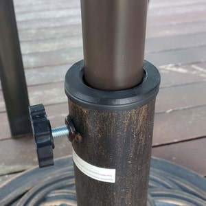 May include: Close-up of a brown metal patio umbrella stand with a black knob and a black plastic ring. The stand is designed to hold an umbrella pole.
