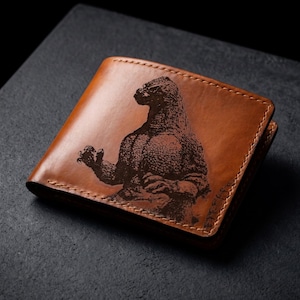 May include: A tan leather bifold wallet featuring a dark brown laser-engraved graphic of a reptilian monster on the front cover. The wallet has visible stitching along the edges and a smooth, polished finish on the exterior surface.