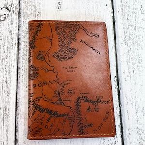 May include: A brown leather wallet featuring a detailed map design. The map includes text such as "Rohan" and "Mordor". The wallet is rectangular and appears to be closed. The background is a white wooden surface.