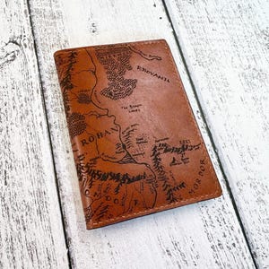 May include: A brown leather passport holder with a detailed map of Middle-earth, including Rohan and Mordor, is displayed on a light wood surface. The map is etched in black, adding contrast to the leather's color. The holder is rectangular.