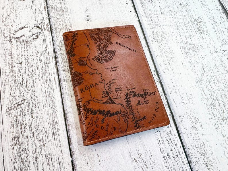 May include: A brown leather passport holder with a detailed map of Middle-earth, including Rohan and Mordor, is displayed on a light wood surface. The map is etched in black, adding contrast to the leather's color. The holder is rectangular.