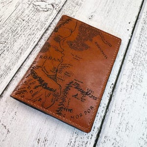 May include: A brown leather passport holder featuring a detailed map design with the words "ROHAN" and "MOR DOR" in black. The passport holder is rectangular and appears to be made of genuine leather.