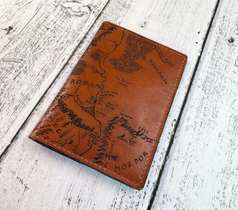 May include: A brown leather passport holder featuring a detailed map design with the words "ROHAN" and "MOR DOR" in black. The passport holder is rectangular and appears to be made of genuine leather.