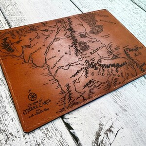 May include: A brown leather wallet featuring a detailed map of Middle Earth. The map includes geographical features, place names, and the text "Middle Earth" and "Map". The wallet is rectangular and appears to be hand-stitched.