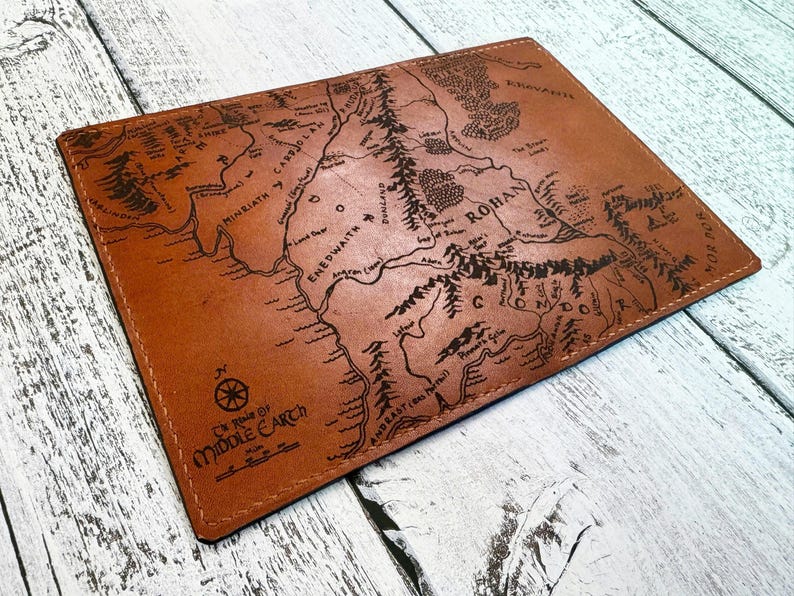 May include: A brown leather wallet featuring a detailed map of Middle Earth. The map includes geographical features, place names, and the text "Middle Earth" and "Map". The wallet is rectangular and appears to be hand-stitched.