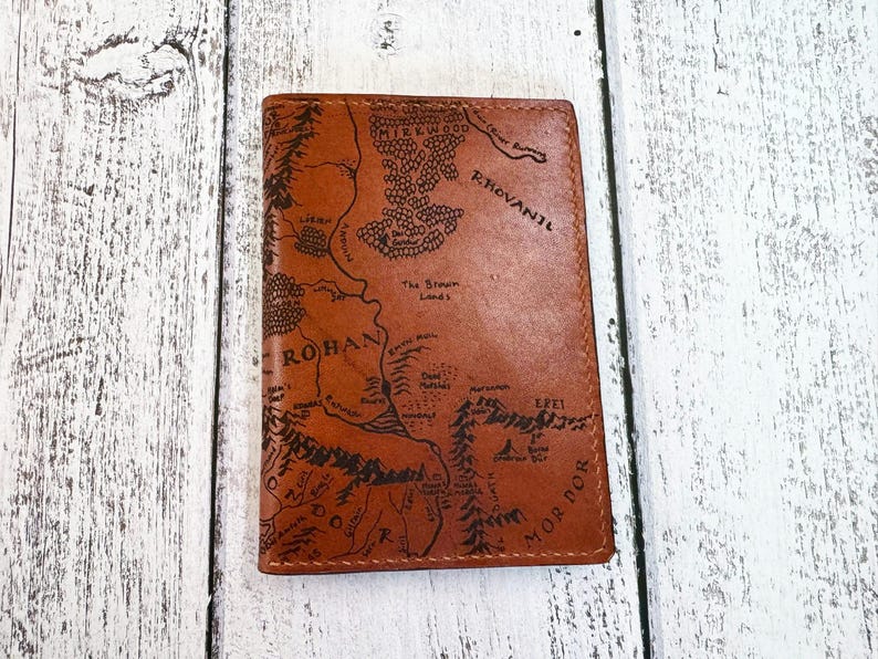 May include: A brown leather wallet featuring a detailed map design. The map includes text such as "Rohan" and "Mordor". The wallet is rectangular and appears to be closed. The background is a white wooden surface.