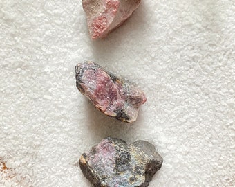 Può includere: Tre pietre preziose grezze, naturali, rosa e nere. La pietra superiore è rosa chiaro, la pietra centrale è rosa scuro con striature nere e la pietra inferiore è grigio scuro con striature rosa e nere.