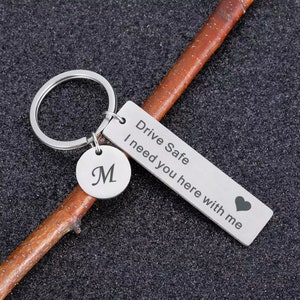 Peut inclure: Un porte-clés argenté avec une étiquette rectangulaire et une étiquette circulaire. L'étiquette rectangulaire porte l'inscription "Drive Safe I need you here with me" et un symbole de cœur. L'étiquette circulaire porte la lettre "M".