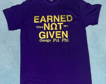 May include: A purple t-shirt with gold text reading "EARNED NOT GIVEN" above "Omega Psi Phi" and "Since 1911". The shirt is made of a soft material and has short sleeves.
