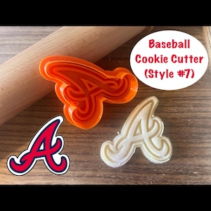 May include: An orange plastic baseball cookie cutter in the shape of the letter "A" sits next to a cookie made with the cutter. A rolling pin is in the background. The text "Baseball Cookie Cutter (Style #7)" is in a white oval.