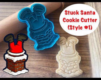 Puede incluir: Un cortador de galletas de plástico azul y un cortador de galletas beige con la forma de Papá Noel atascado en una chimenea. La imagen también incluye una ilustración de dibujos animados de Papá Noel en una chimenea y el texto "Stuck Santa Cookie Cutter (Style #1)".
