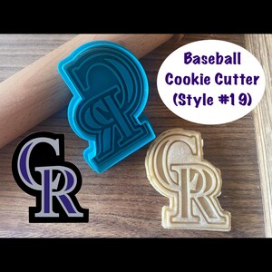 Puede incluir: Un cortador de galletas de béisbol con la forma de las letras "CR" en azul, junto con un gráfico negro y morado del mismo logotipo. También se muestra una galleta horneada con la misma forma. El texto "Baseball Cookie Cutter (Style #19)" está en un óvalo blanco.