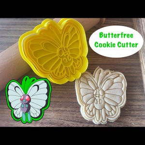 May include: A yellow plastic cookie cutter shaped like a butterfly. The cutter is on a wooden surface next to a cookie that has been cut out using the cutter. The cookie is light brown and has a detailed butterfly design. The text "Butterfree Cookie Cutter" is visible in the image.