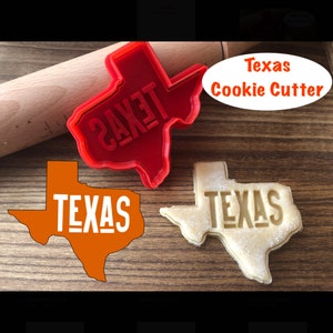 Puede incluir: Un cortador de galletas de plástico rojo en forma de Texas con la palabra "TEXAS" en relieve. El cortador está sobre una superficie de madera junto a un rodillo. Hay dos galletas con forma de Texas, una con la palabra "TEXAS" recortada y otra sin.