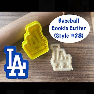 May include: A yellow plastic cookie cutter in the shape of the LA Dodgers logo, with a cookie cut out in the same shape. The text "Baseball Cookie Cutter (Style #28)" is in a blue oval. A wooden rolling pin is in the background.
