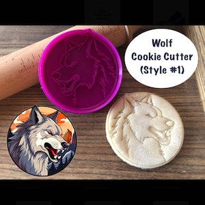 Puede incluir: Un cortador de galletas de lobo de plástico morado con un diseño de cabeza de lobo. El cortador está sobre una superficie de madera con un rodillo y una galleta cortada en forma de cabeza de lobo.