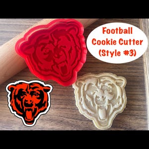 May include: A red plastic cookie cutter in the shape of a bear's head, next to a baked cookie and a sticker with the same design. The text "Football Cookie Cutter (Style #3)" is in a white oval.