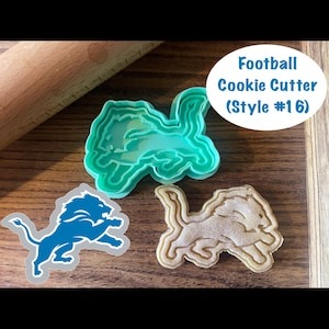 May include: A teal plastic cookie cutter in the shape of a lion, with a blue lion cookie and a tan cookie. The image includes a rolling pin and a white oval with the text "Football Cookie Cutter (Style #16)".