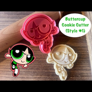 Puede incluir: Un cortador de galletas Buttercup de plástico rojo con la forma del personaje de las Supernenas. La imagen también muestra una galleta terminada con la misma forma y una imagen de dibujos animados de Buttercup. El texto "Buttercup Cookie Cutter (Style #1)" también es visible.