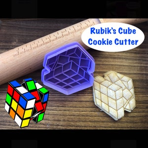 May include: Purple plastic Rubik's Cube cookie cutter on a wooden surface with a rolling pin and a cookie in the shape of a Rubik's Cube. The cookie cutter is labeled "Rubik's Cube Cookie Cutter".