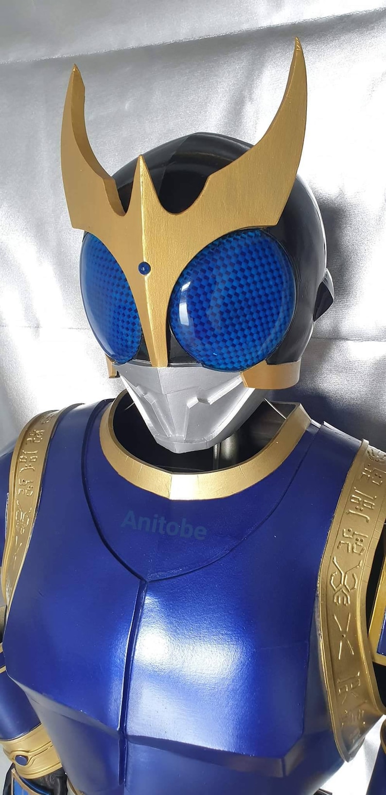 K&ouml;nnte beinhalten: Ein blaues und goldenes Superhelden-Kost&uuml;m mit einem Helm, der eine schwarze Gesichtsmaske und blaue Augen hat. Der Helm hat zwei goldene H&ouml;rner oben. Das Kost&uuml;m hat goldene Akzente und einen goldenen Saum um den Hals.