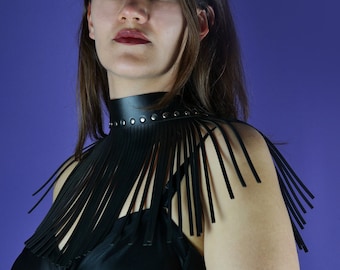 May include: A black leather choker necklace with silver studs and fringe detail. The necklace is worn over a black top against a purple background. The fringe hangs down from the shoulders.