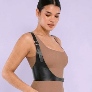 May include: A black leather harness with adjustable straps and a buckle closure. The harness is worn over a nude-colored bodysuit. The design features a shoulder strap and a waist belt, creating a stylish and edgy look.