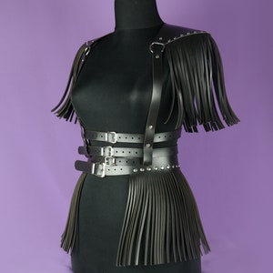 May include: A black leather harness with fringe details and silver buckles. The harness has a wide band around the waist and two straps that go over the shoulders. The fringe is on the shoulders and the bottom of the harness.