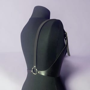 May include: Black leather body harness with adjustable straps and silver-tone hardware. The harness features a simple design with a strap that goes over the shoulder and connects to a waist belt. The harness is displayed on a black mannequin.