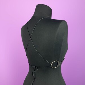 Puede incluir: Arn&eacute;s corporal negro con finas correas que se cruzan en la espalda y un detalle de anillo plateado. El arn&eacute;s se muestra sobre un maniqu&iacute; negro sobre un fondo p&uacute;rpura. Las correas son ajustables y se atan en la espalda.