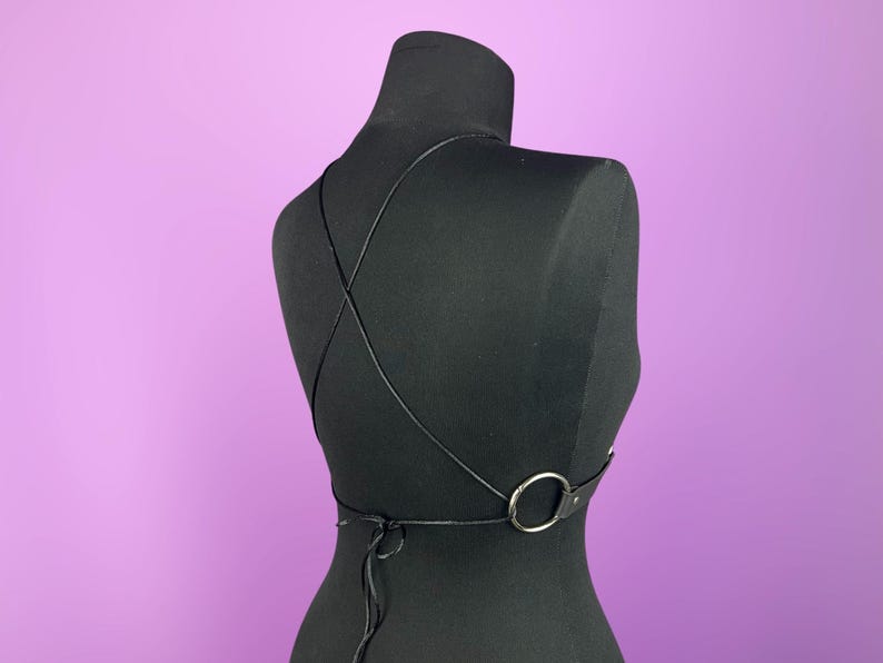 Puede incluir: Arn&eacute;s corporal negro con finas correas que se cruzan en la espalda y un detalle de anillo plateado. El arn&eacute;s se muestra sobre un maniqu&iacute; negro sobre un fondo p&uacute;rpura. Las correas son ajustables y se atan en la espalda.