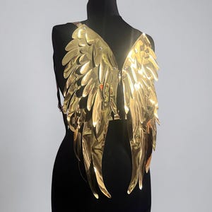 May include: A pair of metallic gold wings constructed from layered, reflective, feather-shaped panels. The wings are attached to a harness system with thin black straps, designed to be worn on the back, featuring a shiny, smooth surface finish.