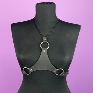 Puede incluir: Arn&eacute;s corporal de cuero negro con tres anillos de metal plateado. El arn&eacute;s se muestra sobre un maniqu&iacute; negro sobre un fondo p&uacute;rpura. El arn&eacute;s tiene un dise&ntilde;o triangular con una correa para el cuello.