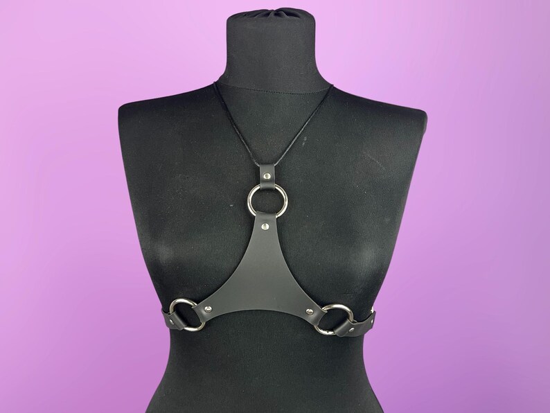 Puede incluir: Arn&eacute;s corporal de cuero negro con tres anillos de metal plateado. El arn&eacute;s se muestra sobre un maniqu&iacute; negro sobre un fondo p&uacute;rpura. El arn&eacute;s tiene un dise&ntilde;o triangular con una correa para el cuello.